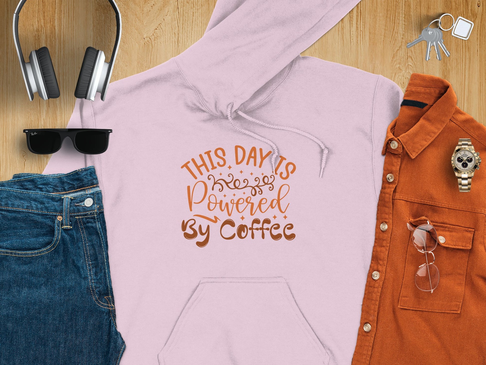 A flat lay features Garment Graphics’ light pink hoodie with This Day is Powered by Coffee text, ideal for coffee lovers. It’s styled with jeans, black sunglasses, large headphones, a rust button-up shirt and glasses, a watch, and keys on wood.