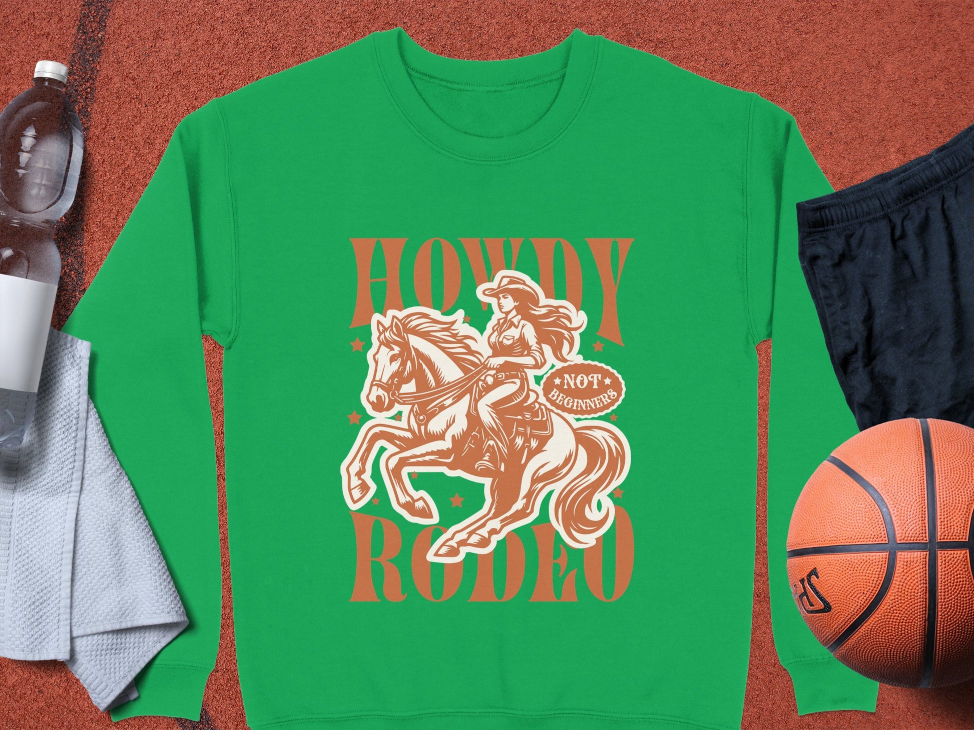 Garment Graphics green graphic print sweatshirt showcases a cowboy on horseback with Howdy Rodeo in brown text and a Not a Beginner banner. Unisex sizing, styled with a towel, water bottle, black shorts, and basketball for sporty flair.