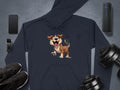 A dark Garment Graphics hoodie with a fun cartoon dog design rests on a gray surface, surrounded by a smartphone, wireless speaker, smartwatch, pair of shoes, and adjustable dumbbell weights—ideal for dog lovers who enjoy playful fashion.