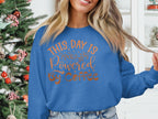 A person in a blue Garment Graphics sweatshirt reading This Day Is Powered by Coffee stands next to a decorated Christmas tree. With long, wavy hair and a slight smile, they playfully hold a strand of hair.