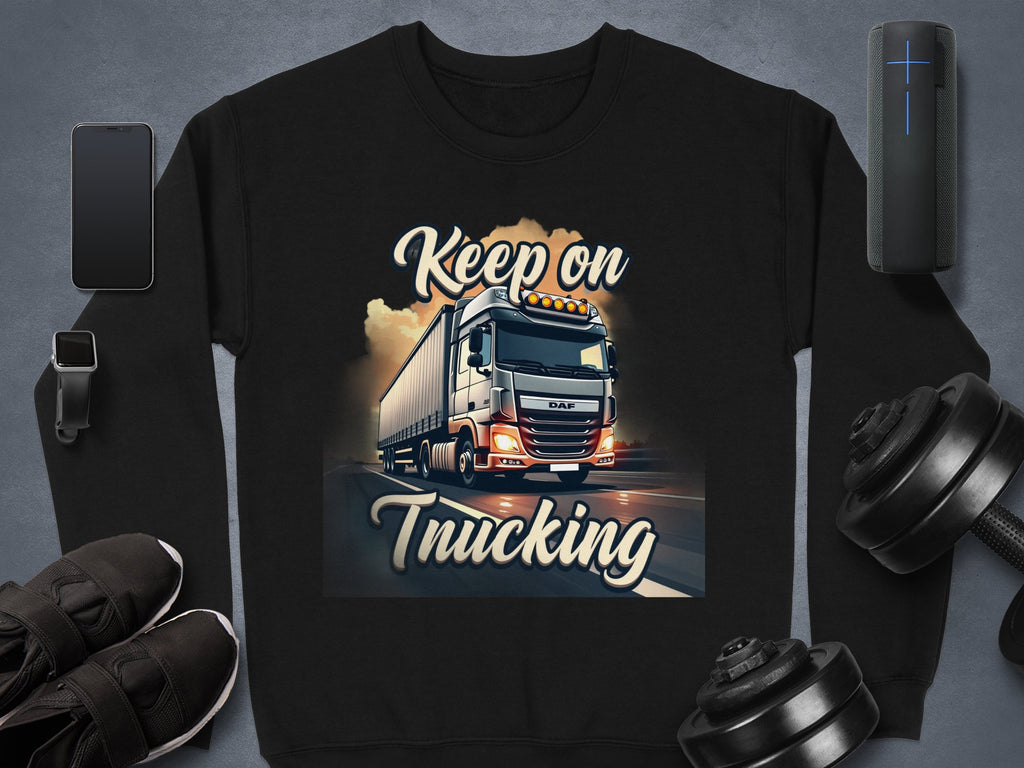 A black Garment Graphics Inspirational Sweatshirt boldly features a truck with Keep on Trucking. Nearby are a smartphone, smartwatch, shoes, portable speaker, and dumbbells on the gray background, evoking adventurous vibes of the open road.