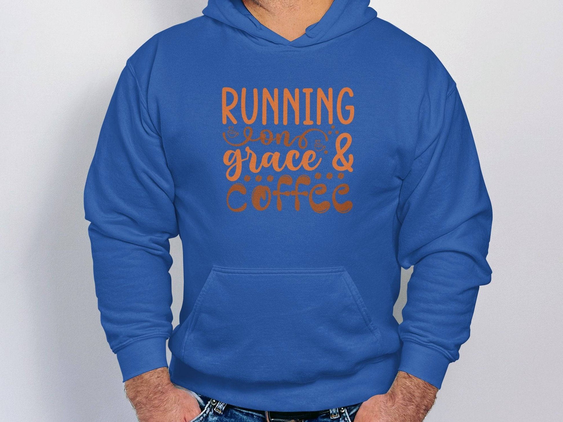 A person is wearing Garment Graphics blue Running on Grace & Coffee hoodie, which features a classic fit and medium-heavy fabric. The stylish orange cursive lettering stands out against the plain white background as they comfortably rest their hands in their pockets.