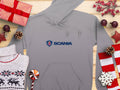 A gray hoodie with the iconic Scania logo from Garment Graphics lies on a wooden surface, surrounded by seasonal décor including wrapped gifts, pinecones, a reindeer-patterned sweater, and a striped candy cane. This unique piece is perfect for festive fashion.