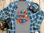 Garment Graphics unisex gray t-shirt features colorful balloons and Happy Birthday text, perfect for celebrations, with a blue plaid shirt, headphones, camera, and takeaway cup on a wooden surface. Ideal for all birthday enthusiasts!.