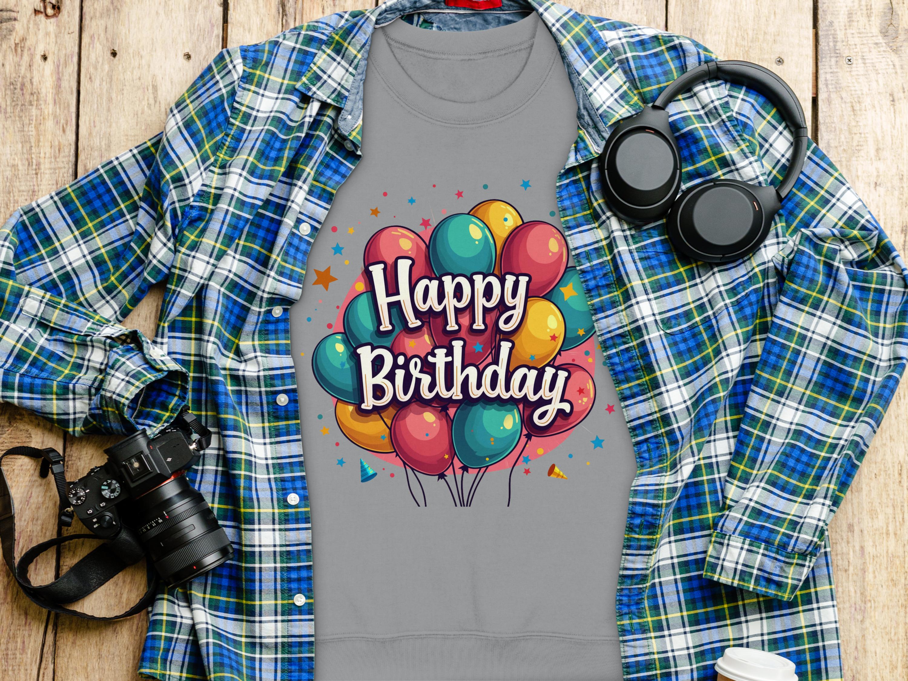 Garment Graphics unisex gray t-shirt features colorful balloons and Happy Birthday text, perfect for celebrations, with a blue plaid shirt, headphones, camera, and takeaway cup on a wooden surface. Ideal for all birthday enthusiasts!.