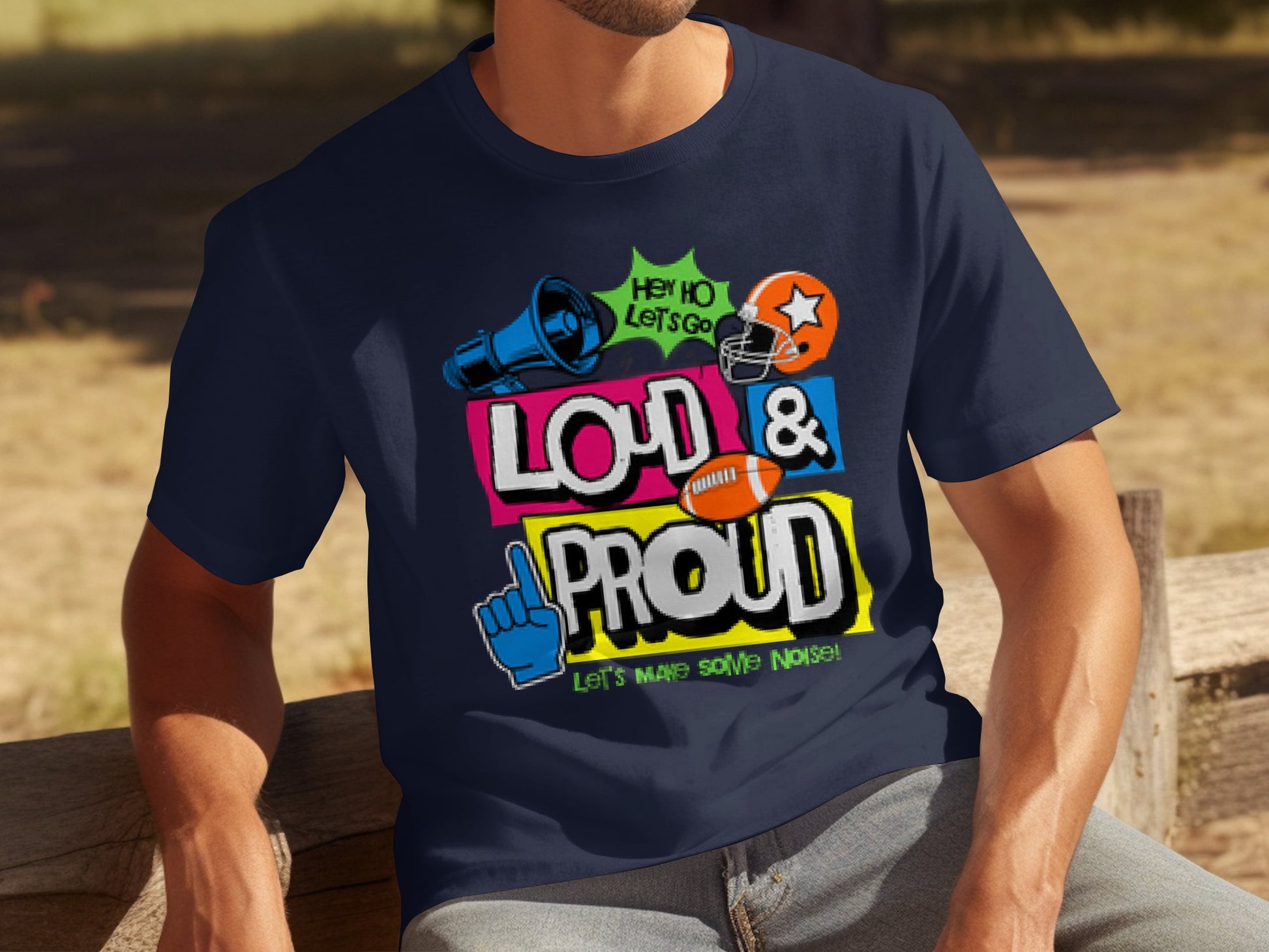 A person sits on a wooden bench wearing a Garment Graphics navy blue football sports t-shirt with a megaphone, foam finger, and helmet, displaying Hey Ho Lets Go! and Loud & Proud Lets Make Some Noise! It captures game passion and is available in family sizing.