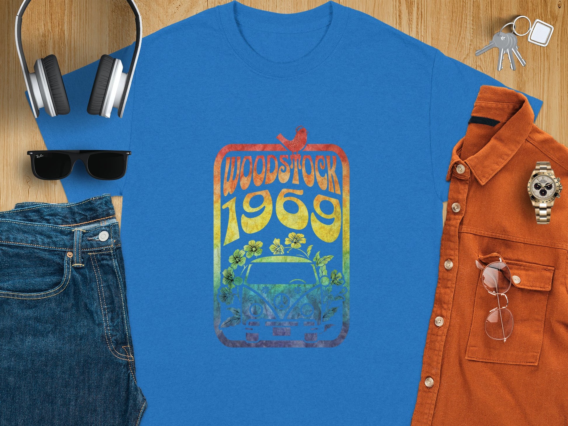 A Garment Graphics blue T-shirt features a retro van, Woodstock 1969, flowers, and a bird. Styled for music lovers, its paired with headphones, sunglasses, jeans, keys, an orange button-up shirt, watch, and glasses on a wooden surface.