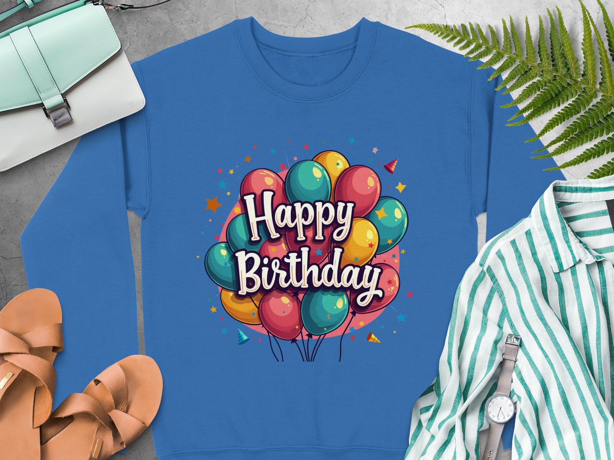 Garment Graphics blue Happy Birthday sweatshirt with a vibrant balloon design is ideal for celebrations. It pairs well with a green-striped shirt, white handbag, brown sandals, and ferns. Available in unisex sizing, its sure to brighten anyones special day.