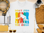 A Garment Graphics white T-shirt showcases a DJ turntable design amid vivid red, yellow, and blue paint strokes. Featuring the slogan Dont Stop the Music, its ideal for music lovers and pairs well with accessories like a scarf, notebook, pen, cup, jacket, sunglasses, and yellow shoes.