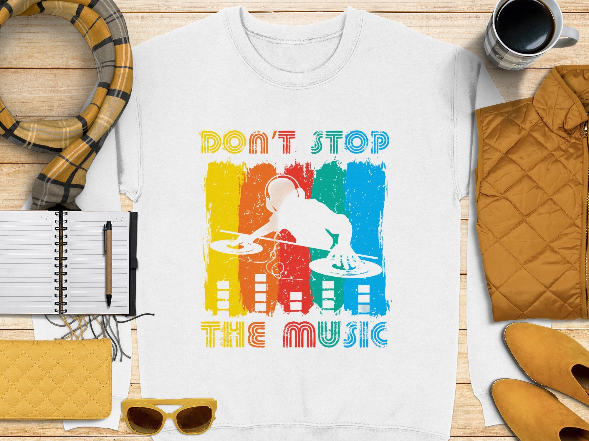 A Garment Graphics white T-shirt showcases a DJ turntable design amid vivid red, yellow, and blue paint strokes. Featuring the slogan Dont Stop the Music, its ideal for music lovers and pairs well with accessories like a scarf, notebook, pen, cup, jacket, sunglasses, and yellow shoes.
