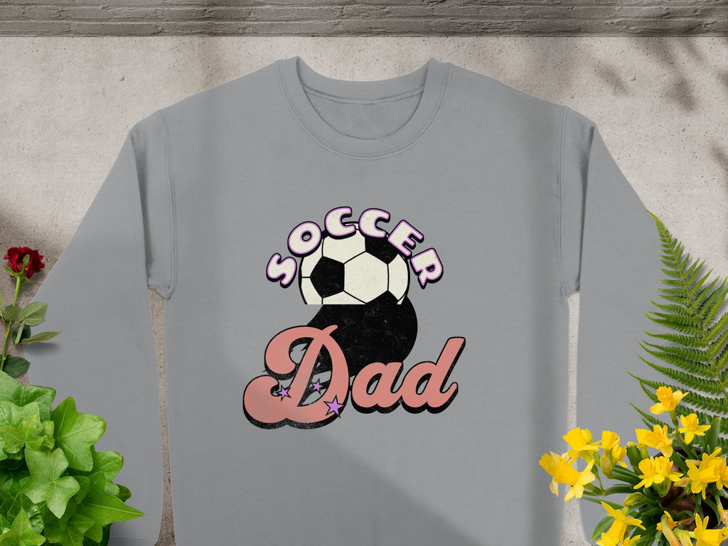 The Garment Graphics gray sweatshirt features a playful retro Soccer Dad graphic, with Soccer above and Dad below a soccer ball. Its displayed on concrete, surrounded by green and yellow plants.