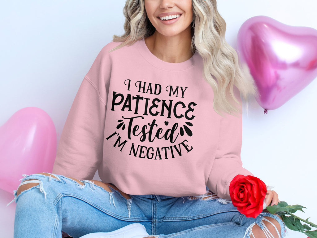 A person with wavy hair sits cross-legged, holding a red rose. Their quirky Garment Graphics sweatshirt says, I had my patience tested Im negative, in unisex sizing. Pink heart-shaped balloons float in the background, adding charm to the casual outing.