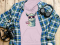 A flat lay displays a light purple Garment Graphics unisex hoodie with a graphic llama in sunglasses and the text No Prob-Llama, surrounded by a blue plaid shirt, headphones, a camera, and a white coffee cup on a wooden surface.