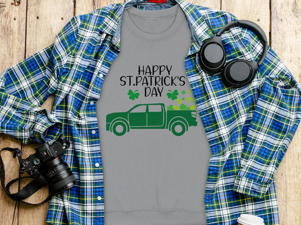 A gray Happy St. Patricks Day Garment Graphics T-shirt featuring a green truck and shamrocks lies flat, surrounded by a blue plaid shirt, headphones, a camera, and a disposable coffee cup on wooden surface.