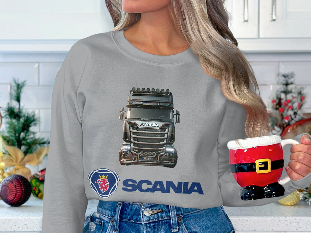 A person in a Garment Graphics gray graphic crewneck sweatshirt featuring a large Scania truck image holds a Santa-themed mug. Holiday decorations, including a small Christmas tree and ornaments, create the perfect festive scene for truck enthusiasts.