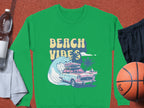 A green Beach Vibes graphic sweatshirt by Garment Graphics features a vintage car with surfboards, waves, and palm trees. In unisex sizing, it complements a water bottle, towel, black shorts, and basketball for retro road trip vibes.