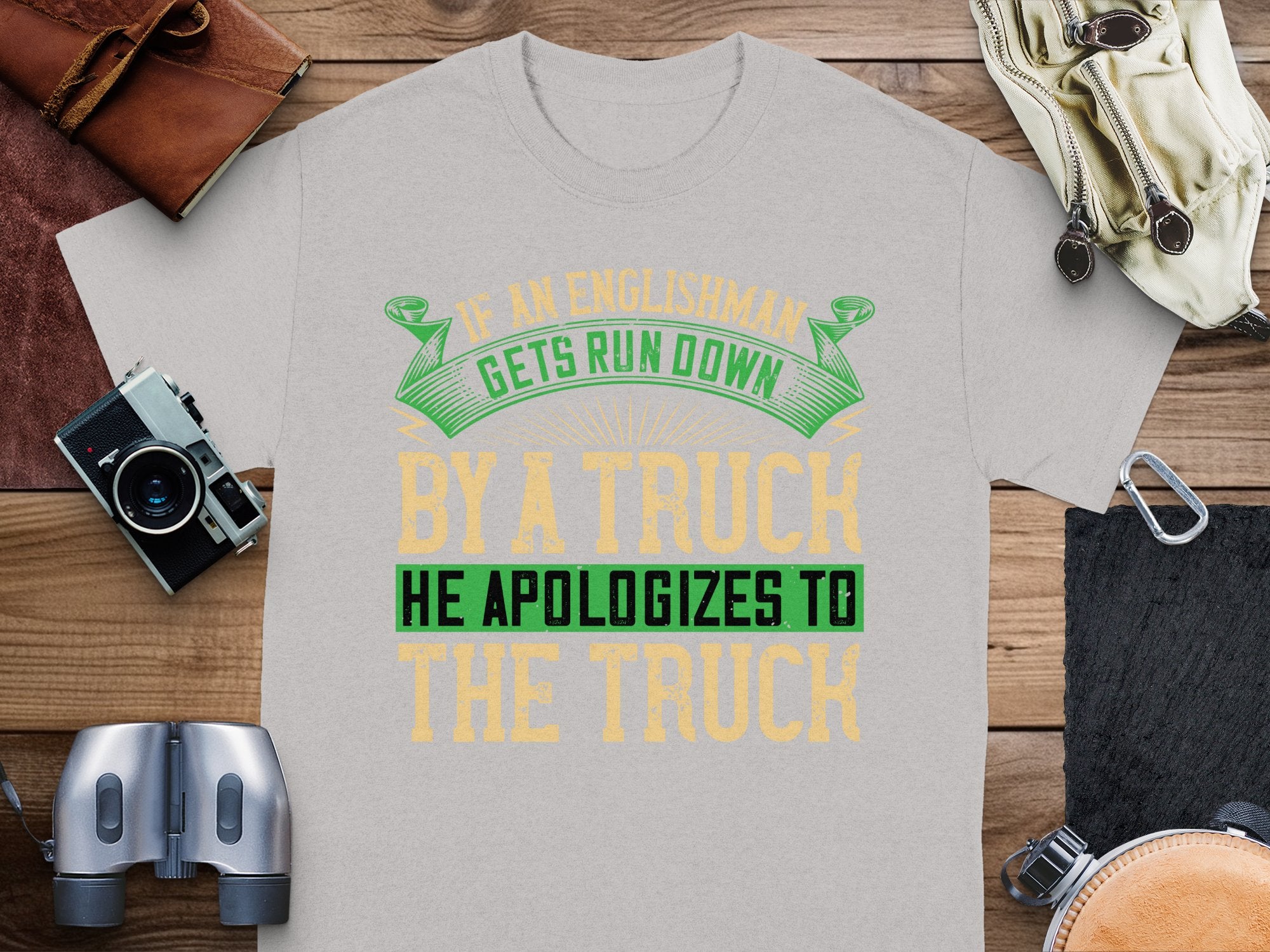 On a wooden surface, Garment Graphics presents a gray t-shirt with the witty green and yellow design, If an Englishman gets run down by a truck, he apologizes to the truck, accompanied by a camera, binoculars, and leather items.