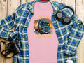 A pink T-shirt by Garment Graphics features a bold truck graphic and the slogan Keep on Trucking, displayed on a wooden surface. Perfect for truck enthusiasts, it’s styled with a blue plaid shirt, headphones, camera, notebook, and coffee cup. A must-have from Graphic T-Shirts collection.