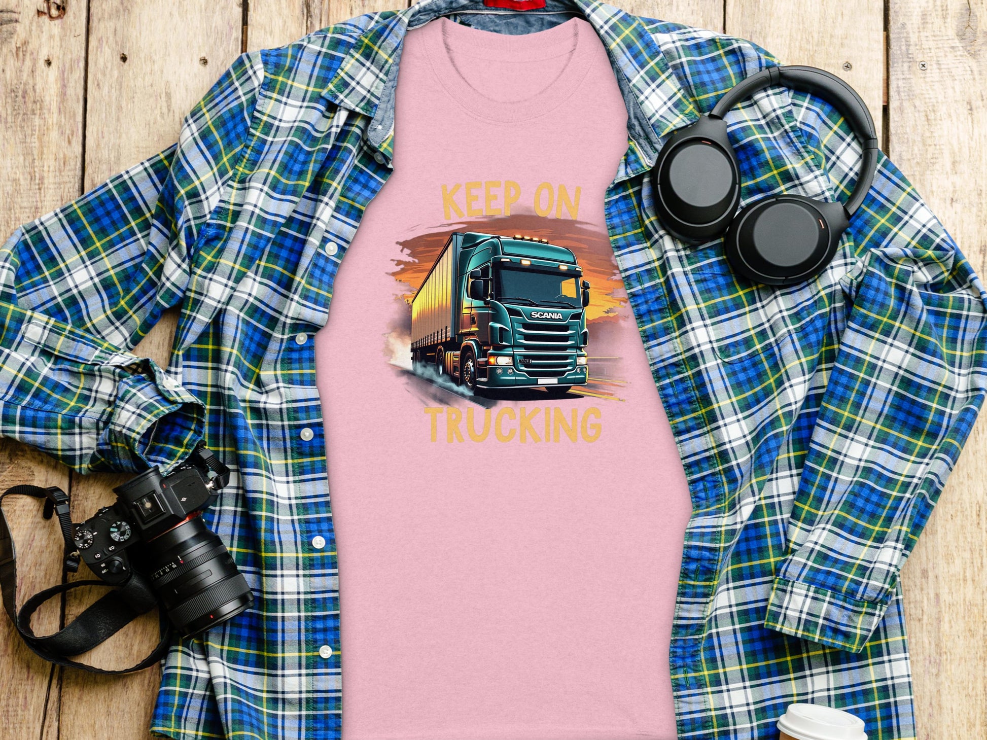A pink T-shirt by Garment Graphics features a bold truck graphic and the slogan Keep on Trucking, displayed on a wooden surface. Perfect for truck enthusiasts, it’s styled with a blue plaid shirt, headphones, camera, notebook, and coffee cup. A must-have from Graphic T-Shirts collection.
