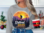 A person holds a Santa-themed mug and wears a Garment Graphics gray retirement sweatshirt featuring a sunset hiker design. The text says, Retired 2025 Not My Problem Anymore. In the background, holiday-decorated kitchen enhances this festive, unisex ensemble.