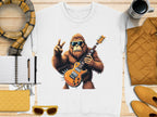 Garment Graphics presents a white sweatshirt with a guitar-playing Bigfoot in sunglasses and a peace sign. Perfect for music lovers, the scene includes cozy details like a yellow quilted jacket, scarf, notebook, pen, clutch, sunglasses, and mug on wood.