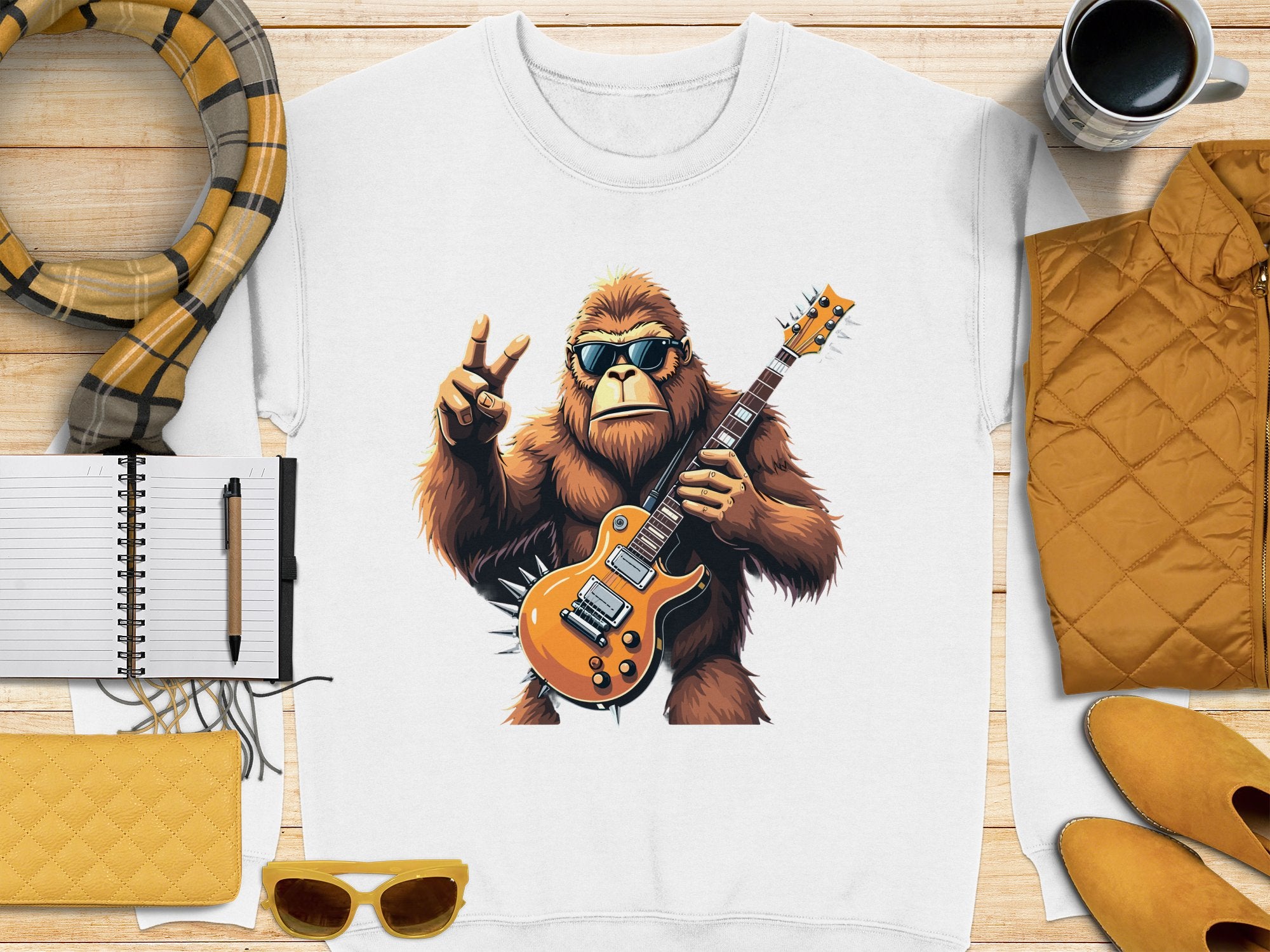 Garment Graphics presents a white sweatshirt with a guitar-playing Bigfoot in sunglasses and a peace sign. Perfect for music lovers, the scene includes cozy details like a yellow quilted jacket, scarf, notebook, pen, clutch, sunglasses, and mug on wood.