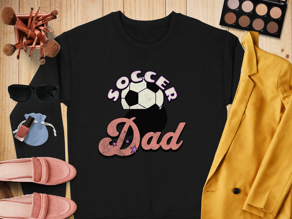 A Garment Graphics retro black Soccer Dad sweatshirt with a soccer ball graphic is laid on a wooden surface, paired with a yellow blazer, pink shoes, sunglasses, makeup brushes, compact palette, nail polish, and blue pouch for an effortlessly chic look.
