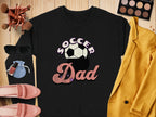 A Garment Graphics retro black Soccer Dad sweatshirt with a soccer ball graphic is laid on a wooden surface, paired with a yellow blazer, pink shoes, sunglasses, makeup brushes, compact palette, nail polish, and blue pouch for an effortlessly chic look.
