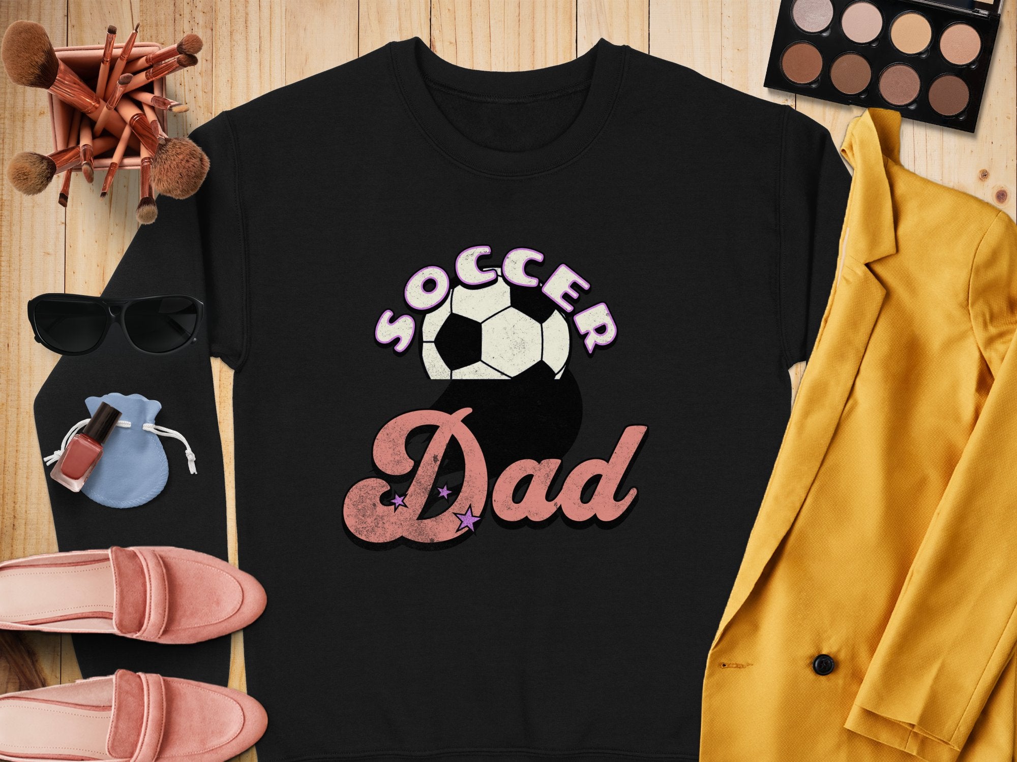 A Garment Graphics retro black Soccer Dad sweatshirt with a soccer ball graphic is laid on a wooden surface, paired with a yellow blazer, pink shoes, sunglasses, makeup brushes, compact palette, nail polish, and blue pouch for an effortlessly chic look.