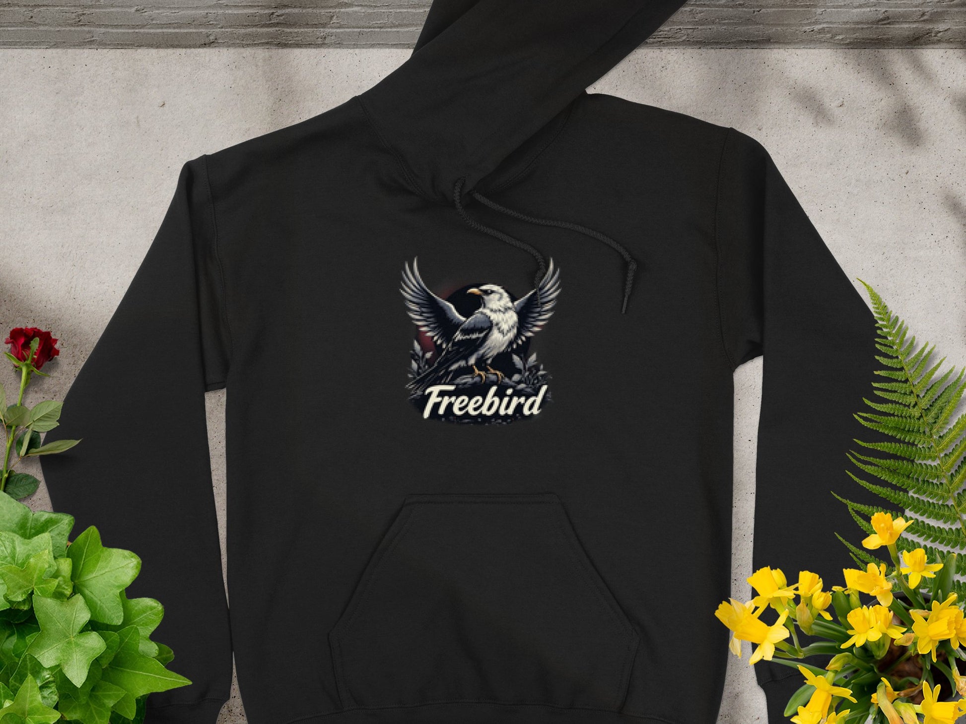 The Garment Graphics Freebird Graphic Pullover Hoodie features a bird with outstretched wings and Freebird beneath. On a concrete background, its artfully surrounded by green leaves, red roses, and yellow flowers, highlighting its distinct style.