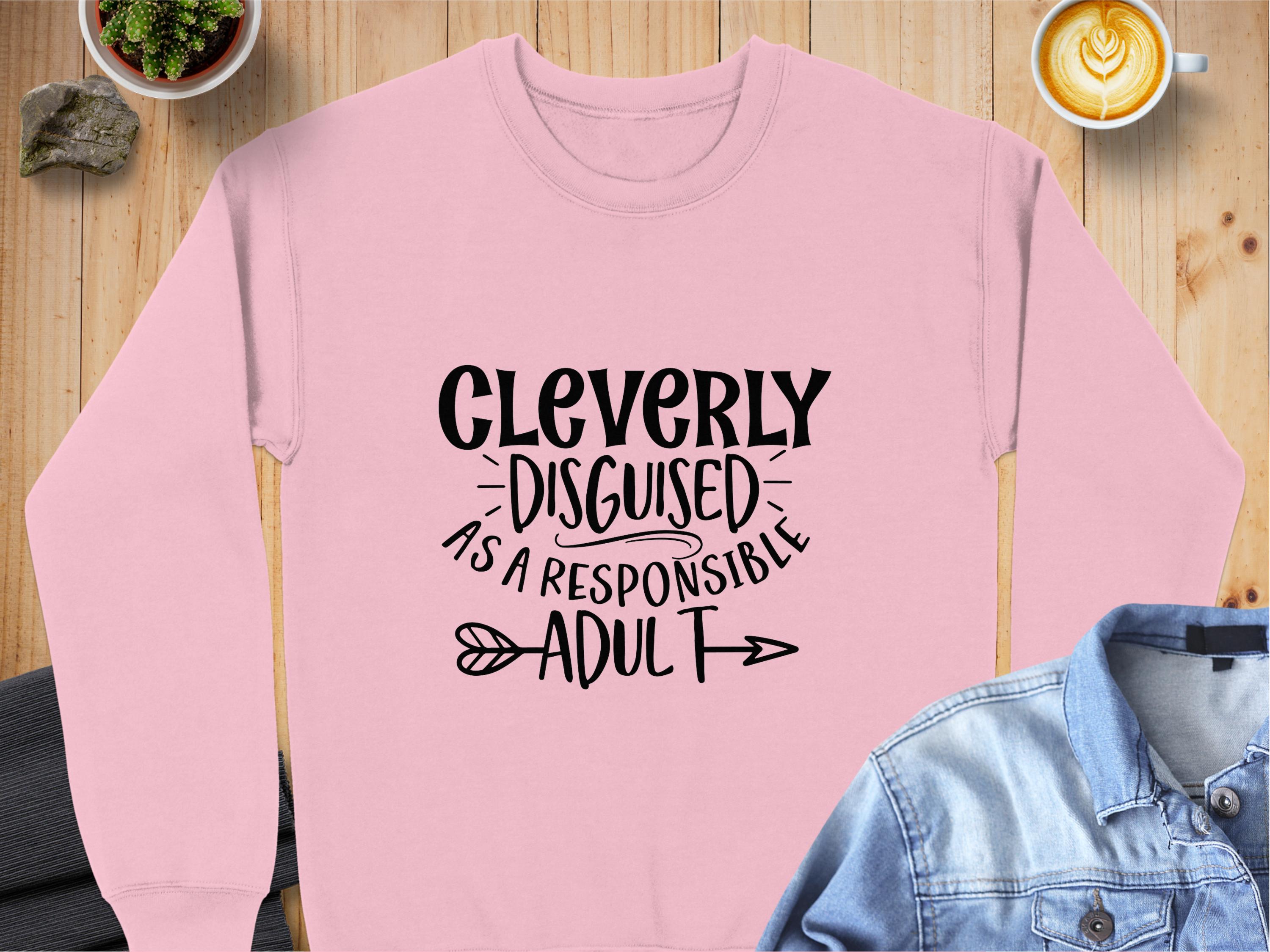 A pink sweatshirt by Garment Graphics with the phrase Cleverly disguised as a responsible adult in bold black letters lies on a wooden table, accompanied by a denim jacket and a latte, offering unisex sizing for this humorous wardrobe essential.