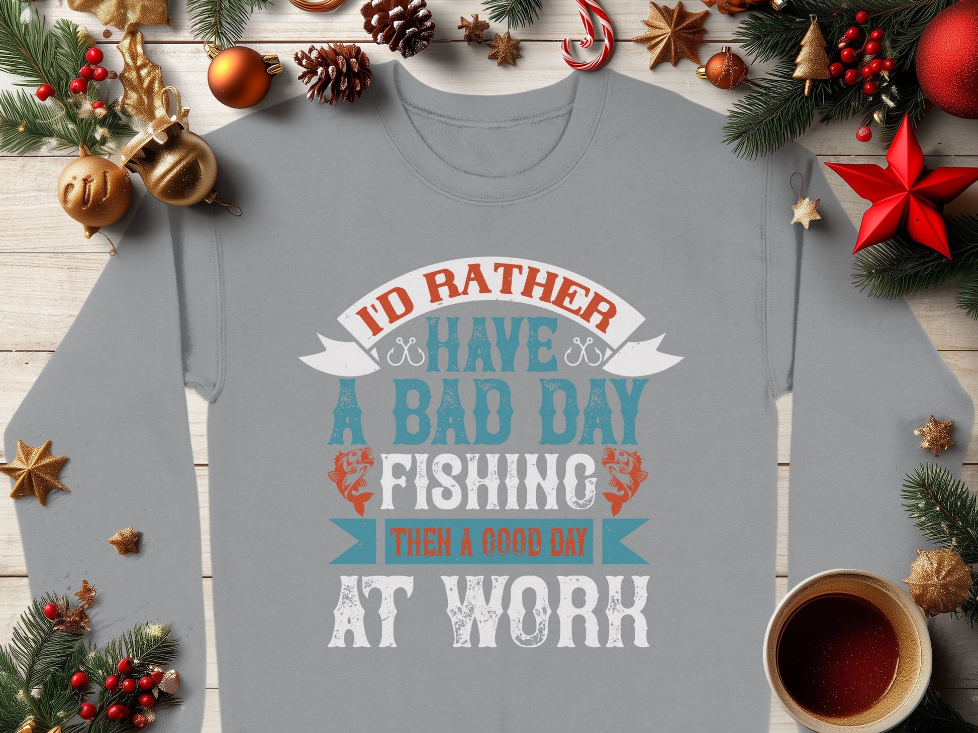 A gray unisex sweatshirt from Garment Graphics, featuring Id rather have a bad day fishing than a good day at work, rests on wood. Surrounded by pine branches, ornaments, dried orange slices, and a red star, it creates a festive vibe for fishing enthusiasts.