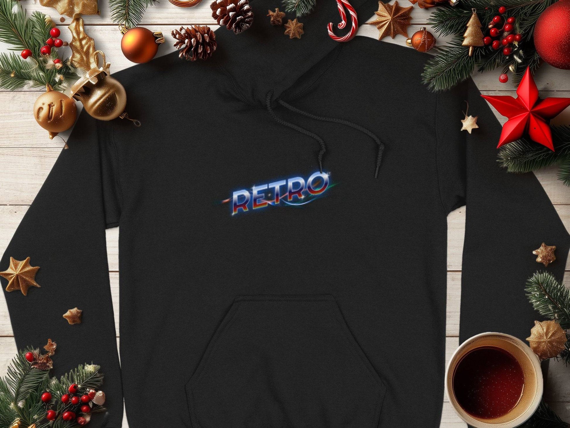 A trendy black hoodie from Garment Graphics, showcasing RETRO in a neon-style font, is set amidst festive decor like colorful pine branches, red and gold ornaments, pinecones, and a cup on a white wooden surface.