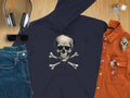 Flat lay featuring Garment Graphics edgy dark hoodie with a skull and crossbones design, paired with blue jeans and a brown shirt. Accessories include headphones, sunglasses, watch, glasses, and keys on a wooden surface.