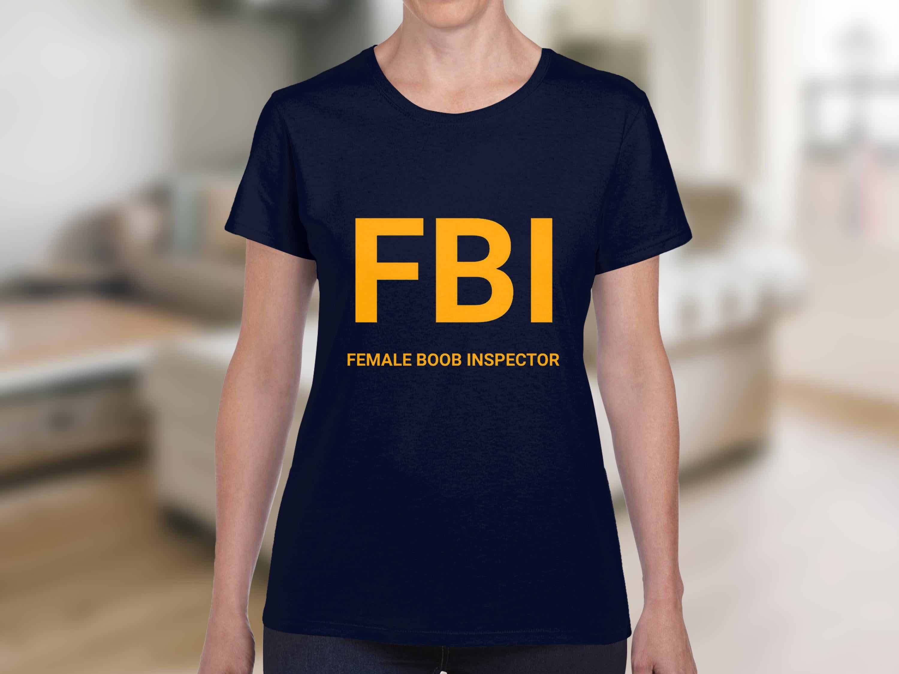 Someone wearing a dark blue Garment Graphics FBI Female Boob Inspector T-shirt makes the perfect gag gift. The bold yellow lettering with smaller text below stands out. Its available in womens sizes for a fun, cheeky fit against a blurred indoor backdrop.