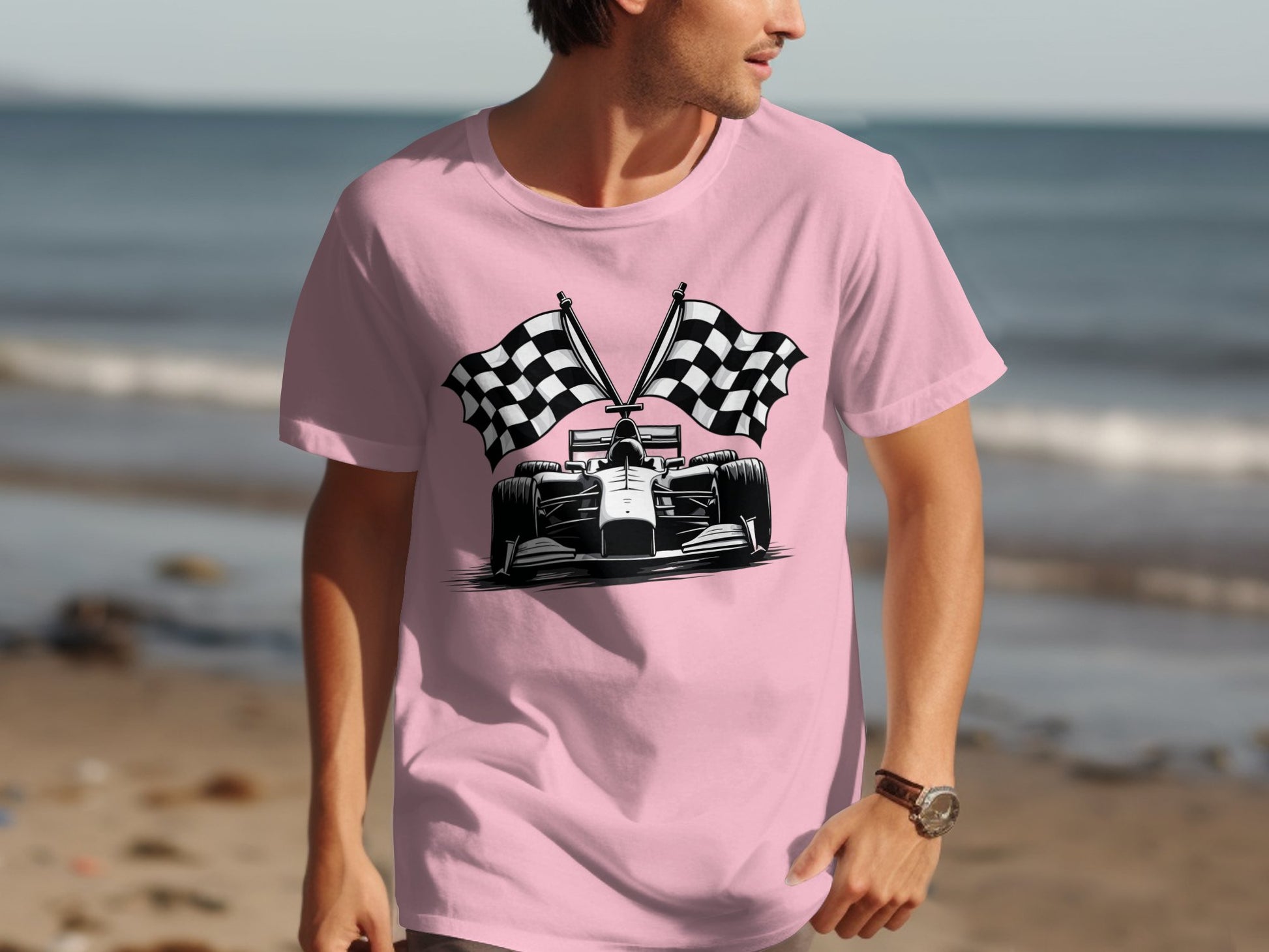 A person in a pink Garment Graphics T-shirt showcasing a motorsport racing car and two checkered flags strolls along the beach, with the ocean visible behind them.