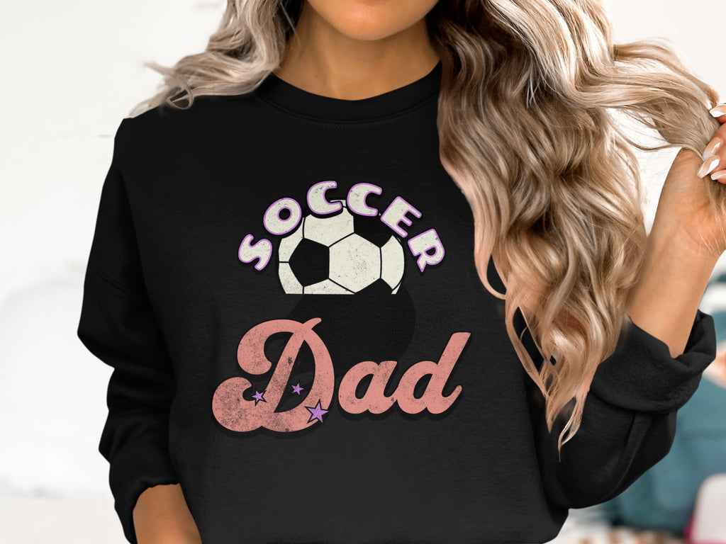 A person with wavy blonde hair wears a Garment Graphics sweatshirt featuring the Soccer Dad design in pink and white letters, complete with a soccer ball graphic above the text.