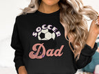 A person with wavy blonde hair wears a Garment Graphics sweatshirt featuring the Soccer Dad design in pink and white letters, complete with a soccer ball graphic above the text.