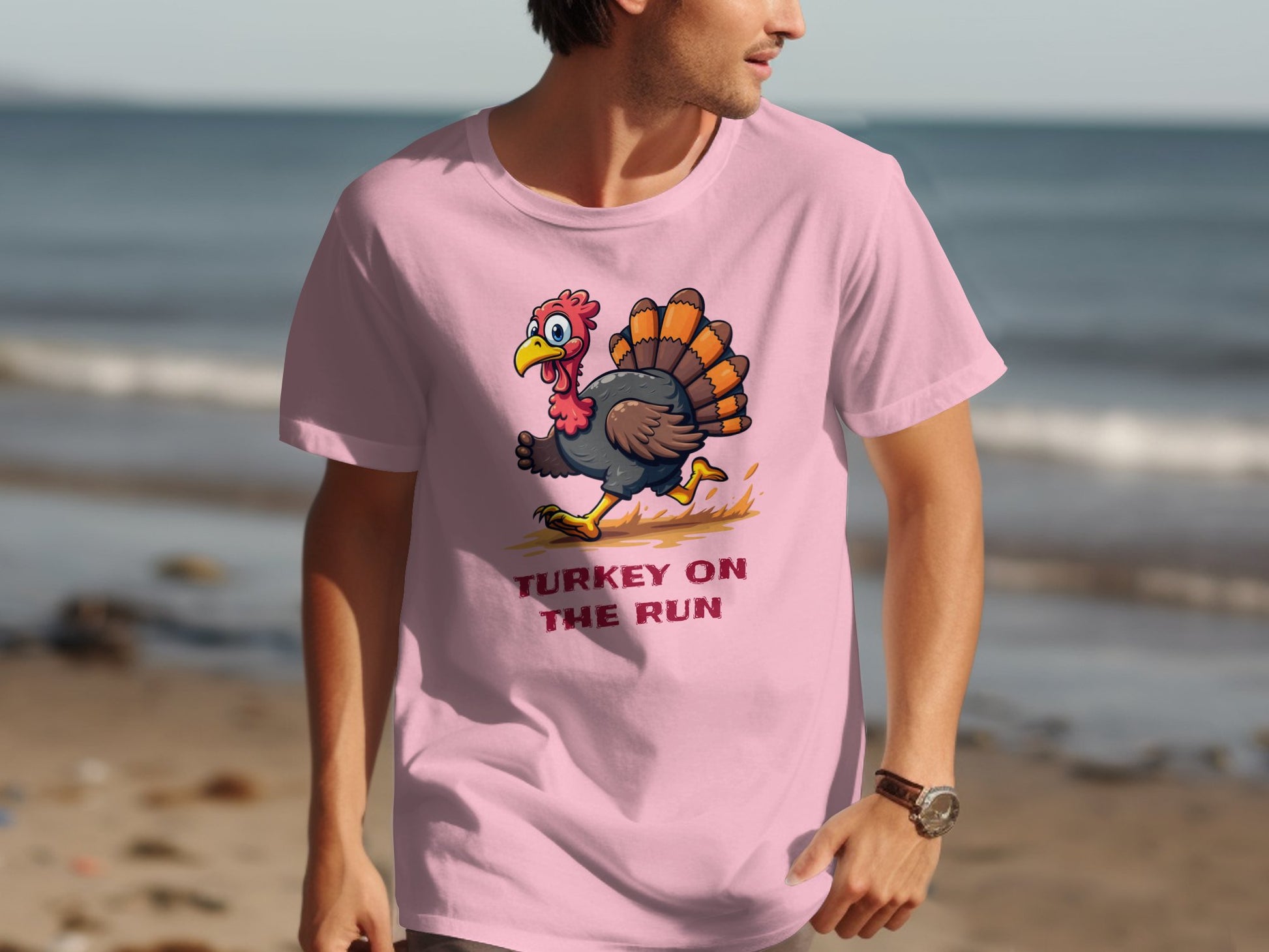 A man walks along the beach in a Garment Graphics classic fit pink T-shirt featuring a humorous turkey cartoon running above the phrase Turkey on the Run, with ocean waves behind him.