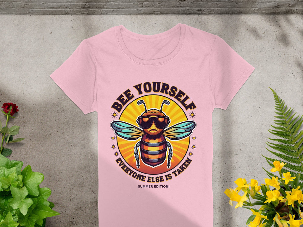 A pink T-shirt from Garment Graphics lies on a floral-adorned concrete surface. It features the Bee Yourself: Summer Edition design with a sunglass-wearing bee in a sunburst, tailored for women and reminding you to be yourself since everyone else is taken!.