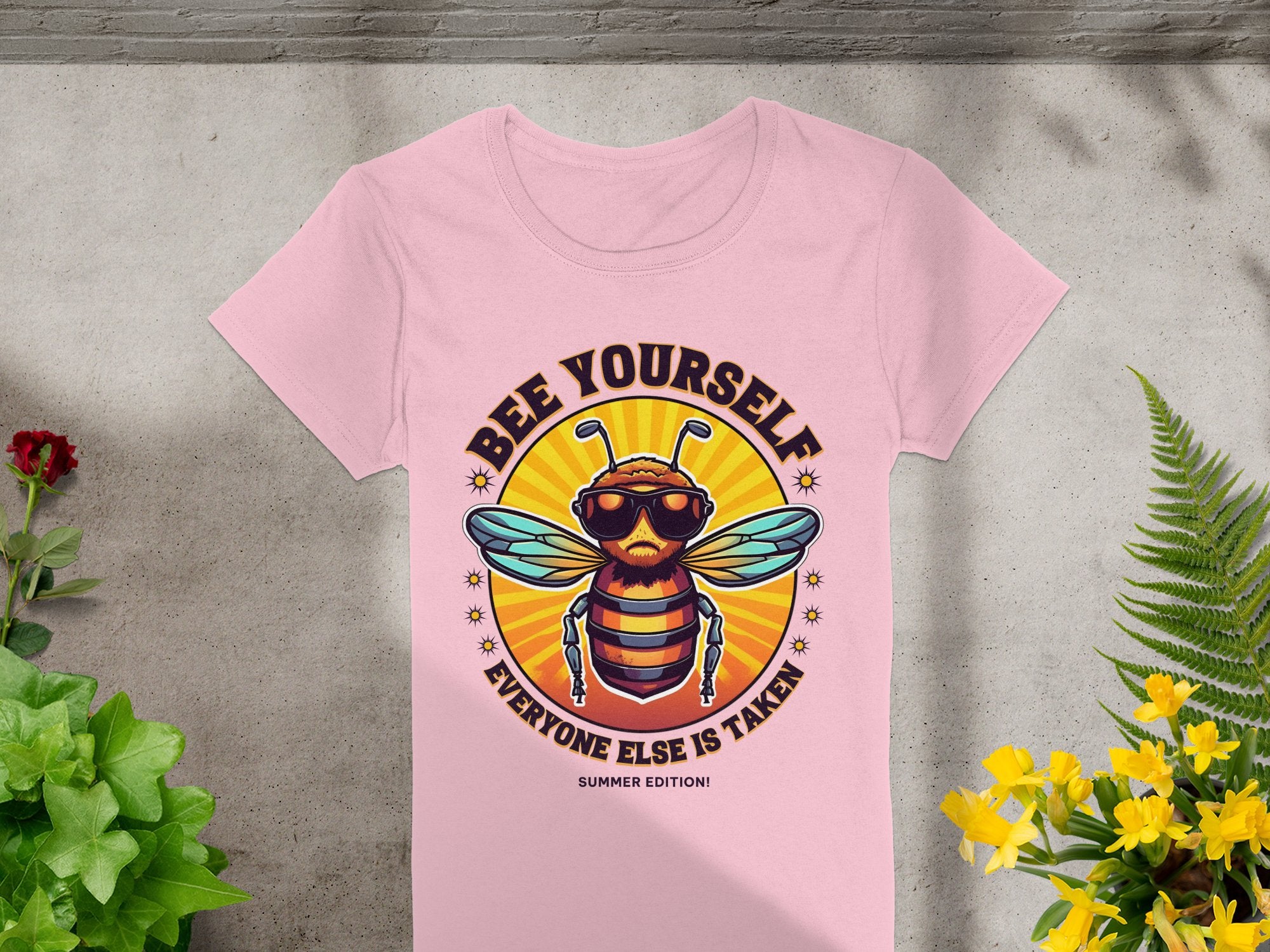 A pink T-shirt from Garment Graphics lies on a floral-adorned concrete surface. It features the Bee Yourself: Summer Edition design with a sunglass-wearing bee in a sunburst, tailored for women and reminding you to be yourself since everyone else is taken!.