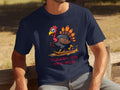A person sits outdoors on a wooden bench in a navy Garment Graphics T-shirt featuring a funny sprinting turkey cartoon with Turkey on the Run text. The blurred background reveals trees and grass.