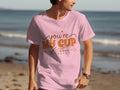 A man on a beach wears a 100% cotton graphic T-shirt from Garment Graphics, featuring the phrase youre my cup of tea in charming brown and orange lettering. The tranquil ocean and soft sand provide the perfect backdrop for his stylish statement.