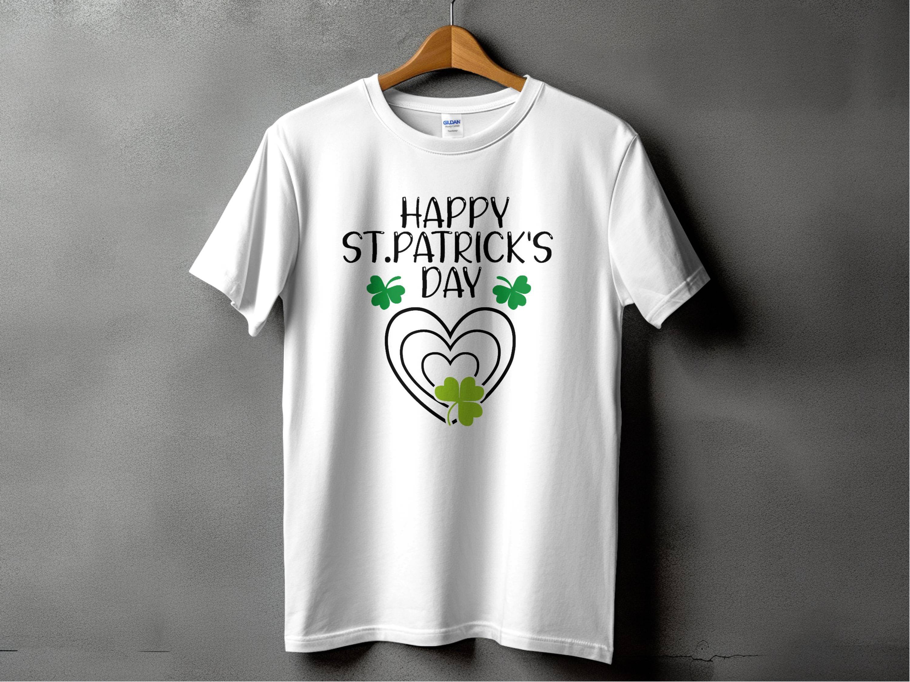 Garment Graphics offers a festive graphic T-shirt featuring a Happy St. Patricks Day design with a heart and shamrocks. It hangs stylishly on a wooden hanger against a gray backdrop, ready for celebration in style.