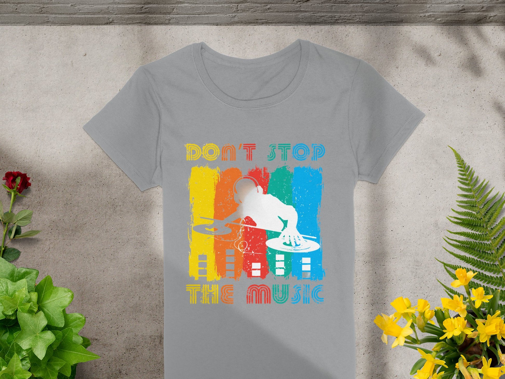The Garment Graphics DJ tee showcases a vivid design of a DJ with turntables, ideal for music lovers. Bright stripes in red, yellow, blue, and orange dominate the background with Dont Stop the Music, set against concrete and surrounded by plants, embodying dynamic style.