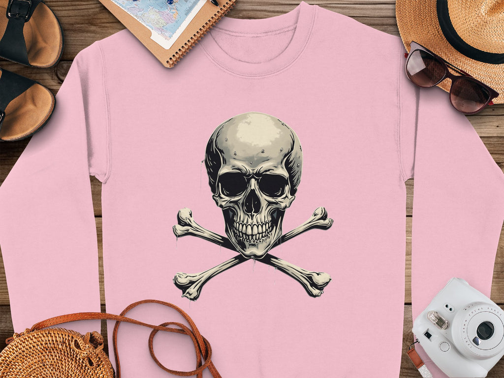 A pink unisex sweatshirt from Garment Graphics, featuring a skull and crossbones design, is on a wooden surface. Its surrounded by sandals, sunglasses on a straw hat, a notebook, and a camera—all creating a carefree vacation atmosphere.