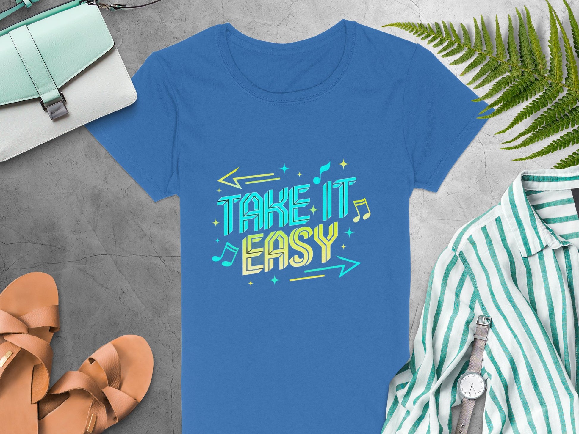 A Garment Graphics Take It Easy T-shirt in blue features vibrant graphics and a playful font with arrows and musical notes, creating a stylish contrast on a gray background. It pairs well with brown sandals, a striped green shirt, watch, mint bag, and green leaves.