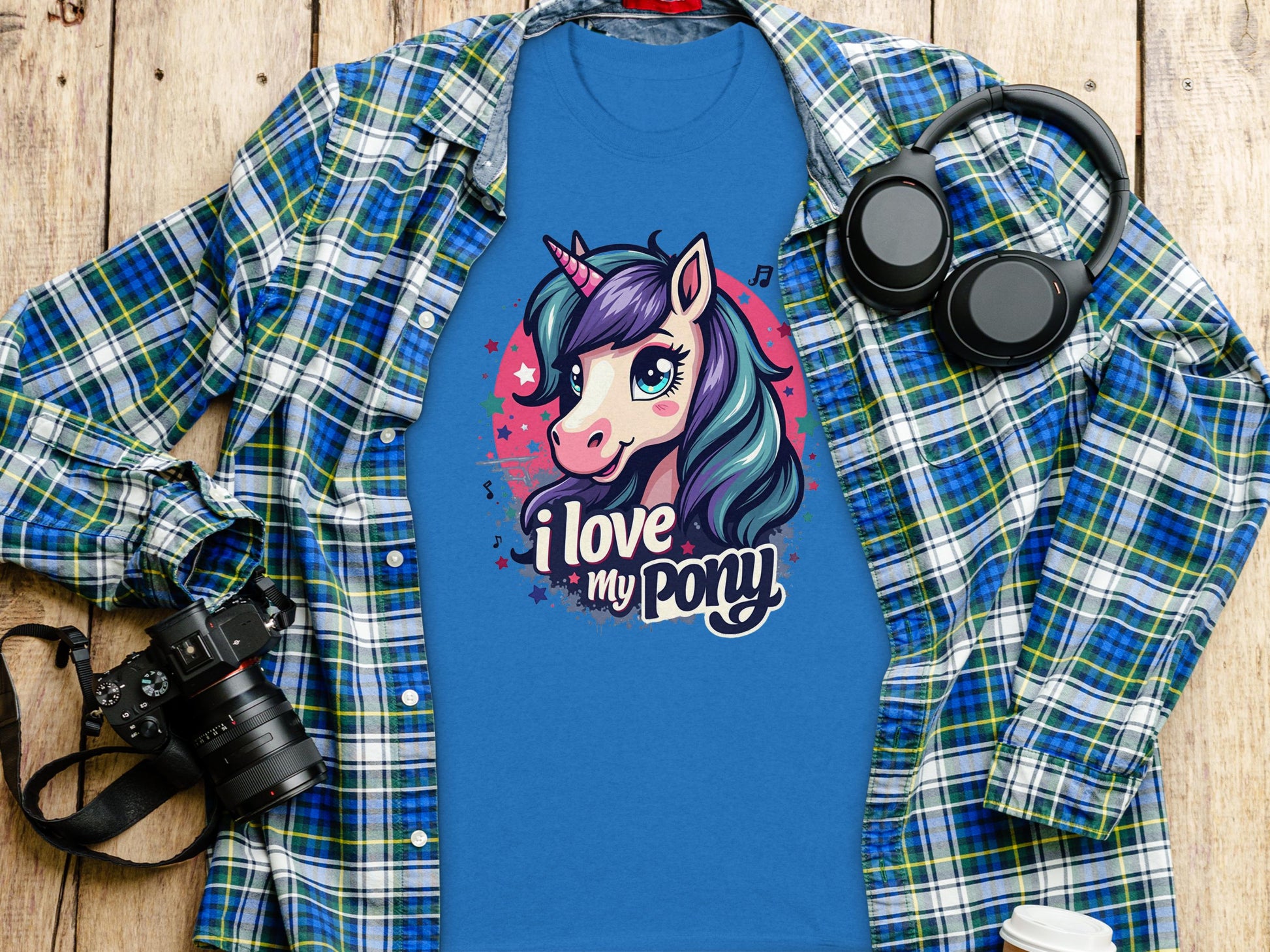 A Unicorn enthusiasts dream, Garment Graphics’ blue t-shirt showcases a pony graphic with I love my pony. It’s styled on a wood surface with a plaid shirt, alongside a camera, headphones, white cup, and notebook.