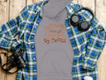 A chic Garment Graphics gray hoodie with This Day is Powered by Coffee lies on a wooden surface, perfect for coffee lovers. Surrounded by a blue-green plaid shirt, headphones, camera, and takeout coffee cup, its a true tribute to stylish Coffee Hoodies.