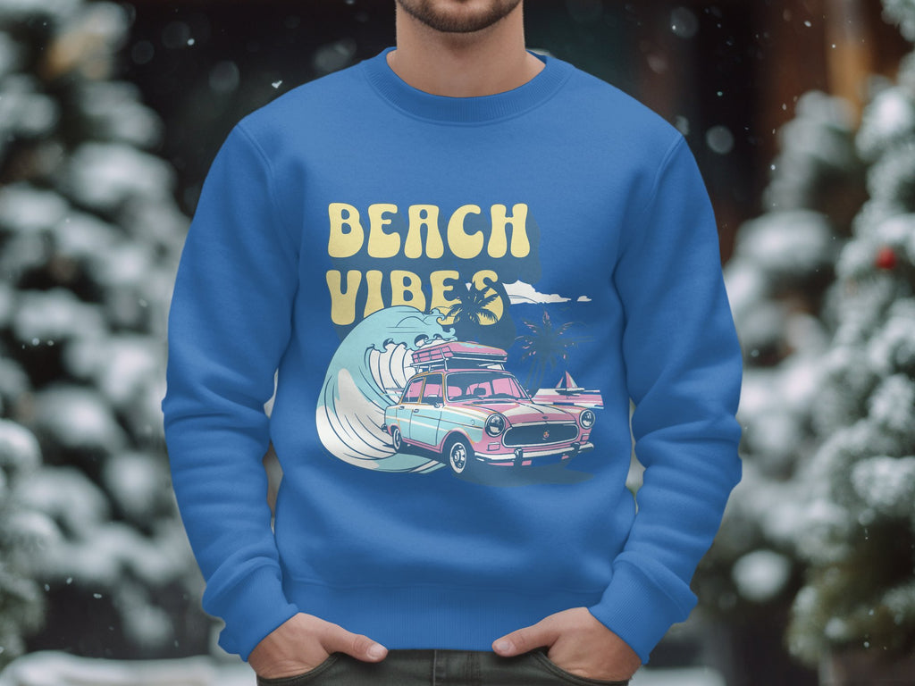 A person wears a blue Garment Graphics Beach Vibes sweatshirt with a pink car, surfboard, and wave design. The unisex sizing suits anyone. Snow-covered trees in the background add retro road trip vibes to the winter scene.